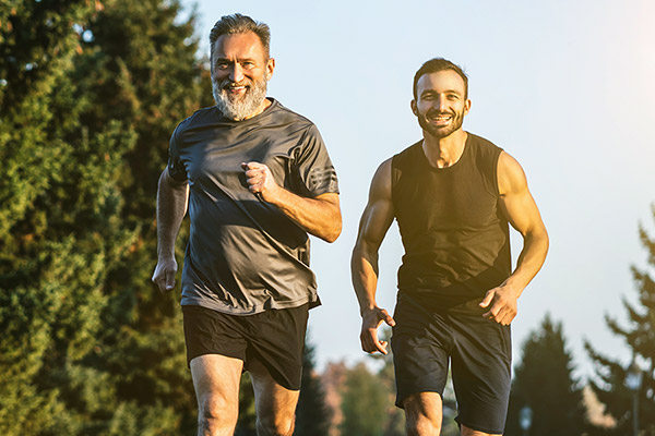 Two middle aged white men jog on a path through trees while smiling. 